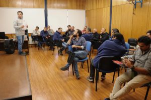 Funcionario de pie, frente a otros funcionarios (hombres) sentados en grupo mirando la pantalla.